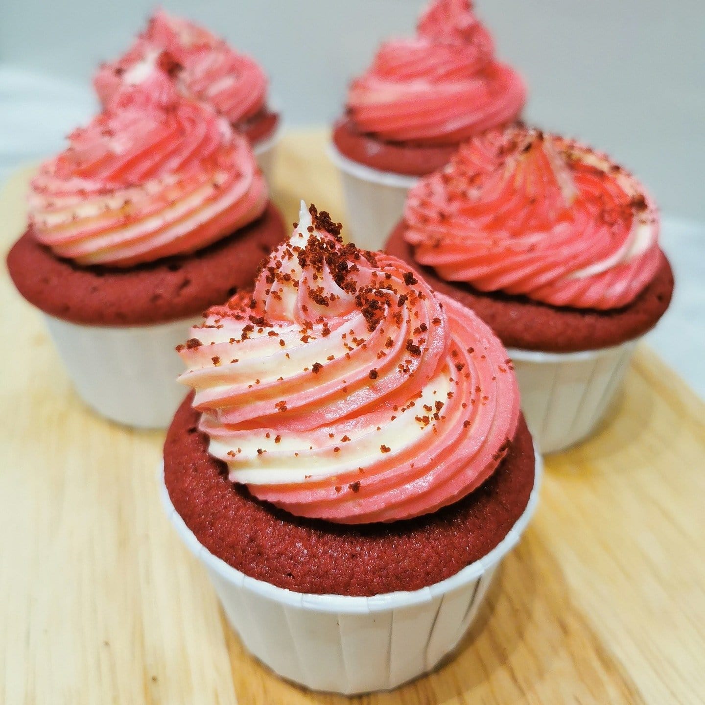 5 petits cupcakes rouges dans de petits papiers blancs avec un glaçage blanc et rose, le tout sur une planche en bois