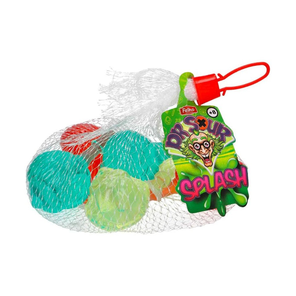 Un emballage en filet qui permet de voir des boules transparentes contenant des liquides de couleurs différentes ; jaune, rouge, bleu et une étiquette verte sur le côté droit avec un visage de Dr aux cheveux verts. Le tout sur fond blanc