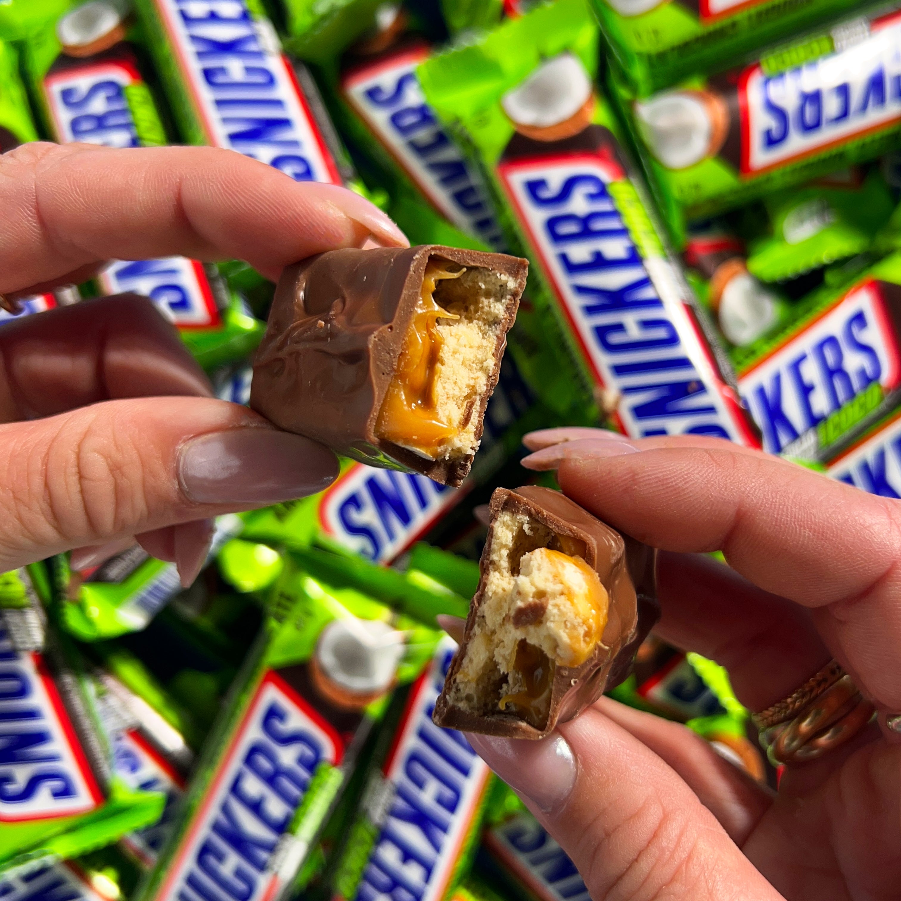 Une barre chocolatée coupée en 2 et tenue par 2 mains, à l'arrière il y a plein d'emballages verts de Snickers Coco