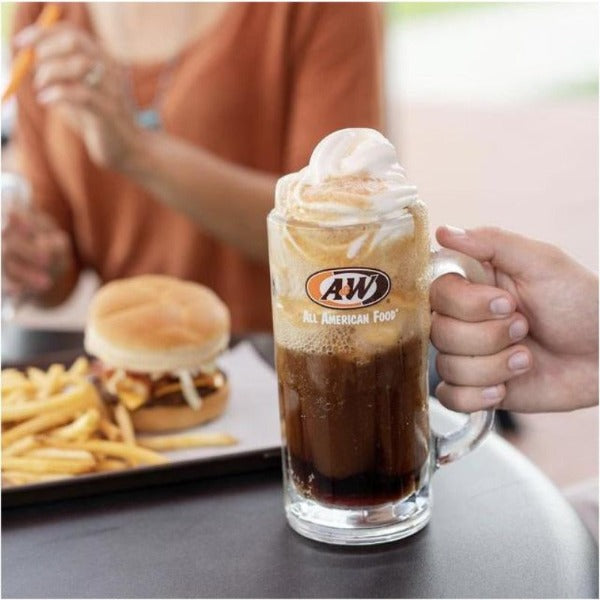 Une choppe de bière avec le logo A&W et une main qui la tient, à l’arrière un plateau avec un hamburger et des frites et une femme avec une blouse orange 