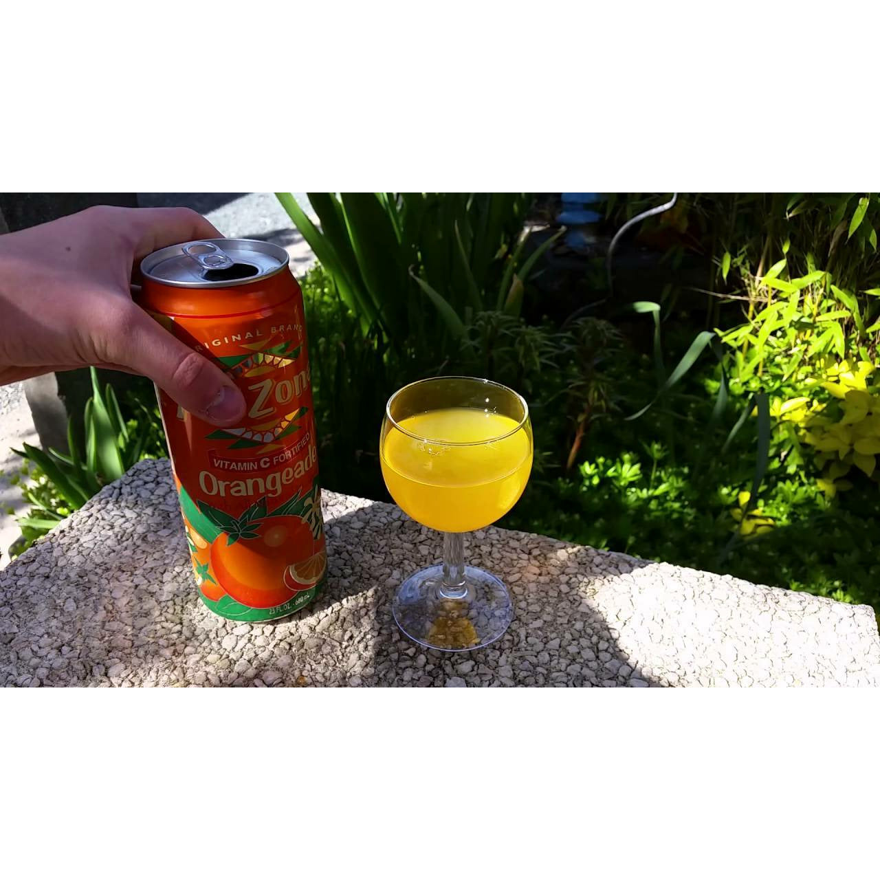Une main qui tient une canette orange ouverte et à droite un verre de boisson sur une dalle en pierre et devant des plantes