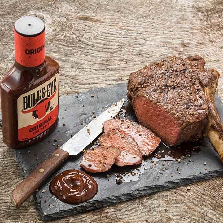 Une grosse viande à l’os avec un couteau sur une planche noir et sur le côté gauche une bouteille brune avec une tête de taureau rouge. Le tout sur une table en bois brut 