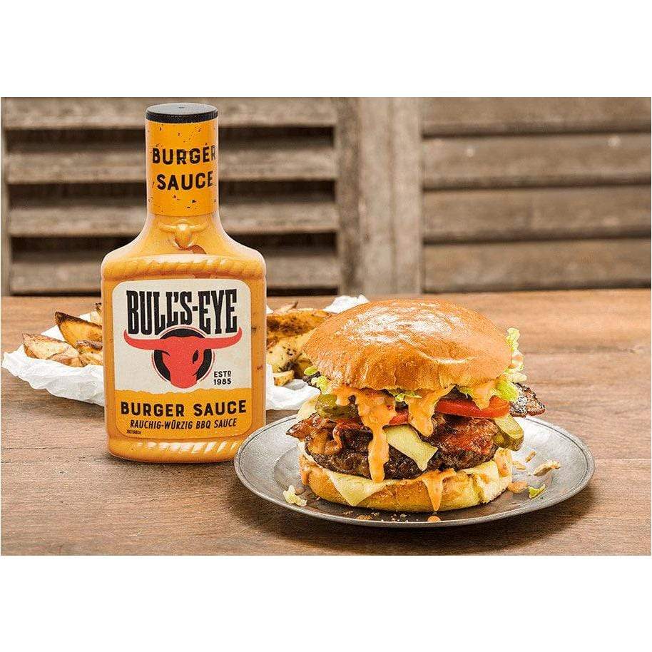 Une bouteille orange avec une tête de taureau rouge au centre, à droite un hamburger dans une assiette grise et derrière une assiette de frites maison. Le tout sur une table en bois