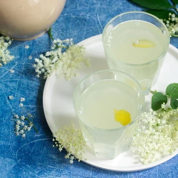 Deux verres d’une boisson jaune pâle sur une assiette blanche avec des fleurs de sureau. Le tout sur une table avec une nappe bleue
