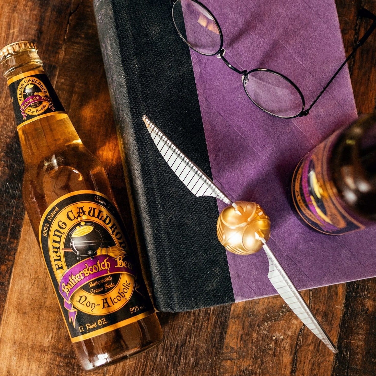 A gauche une bouteille en verre remplie d’une boisson brune, à droite un livre mauve, des lunettes et le Vif d’or. Le tout sur une table en bois