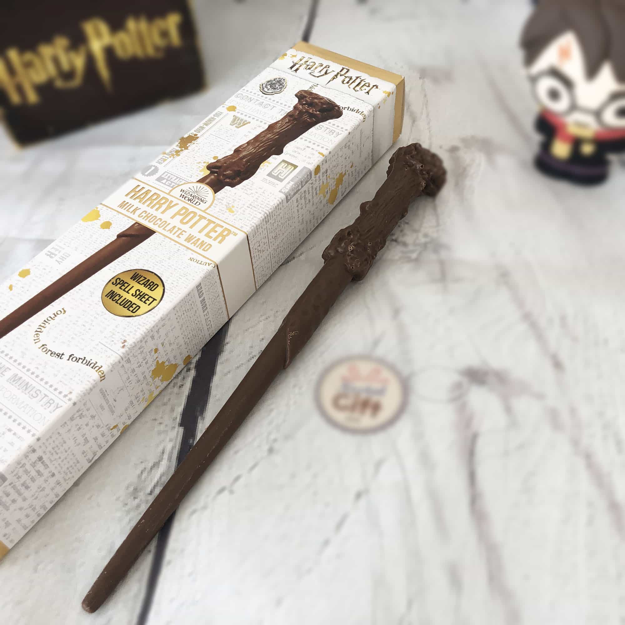 Un emballage blanc avec sur toute sa longueur la baguette magique d’Harry Potter en chocolat et à droite la baguette en chocolat. Le tout sur une table à nappe blanche