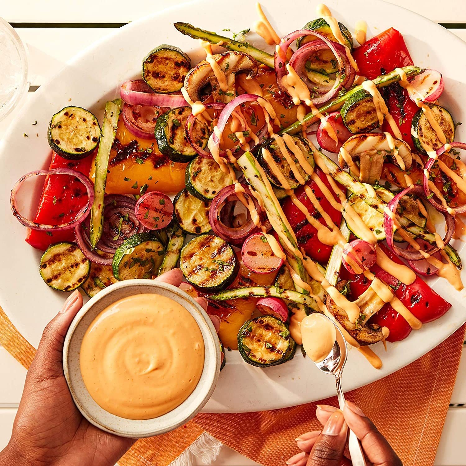 Un plat blanc rempli de légumes avec une sauce couleur pêche au dessus, il y a 2 mains qui tiennent à gauche un bol de sauce et à droite une cuillère avec un peu de sauce