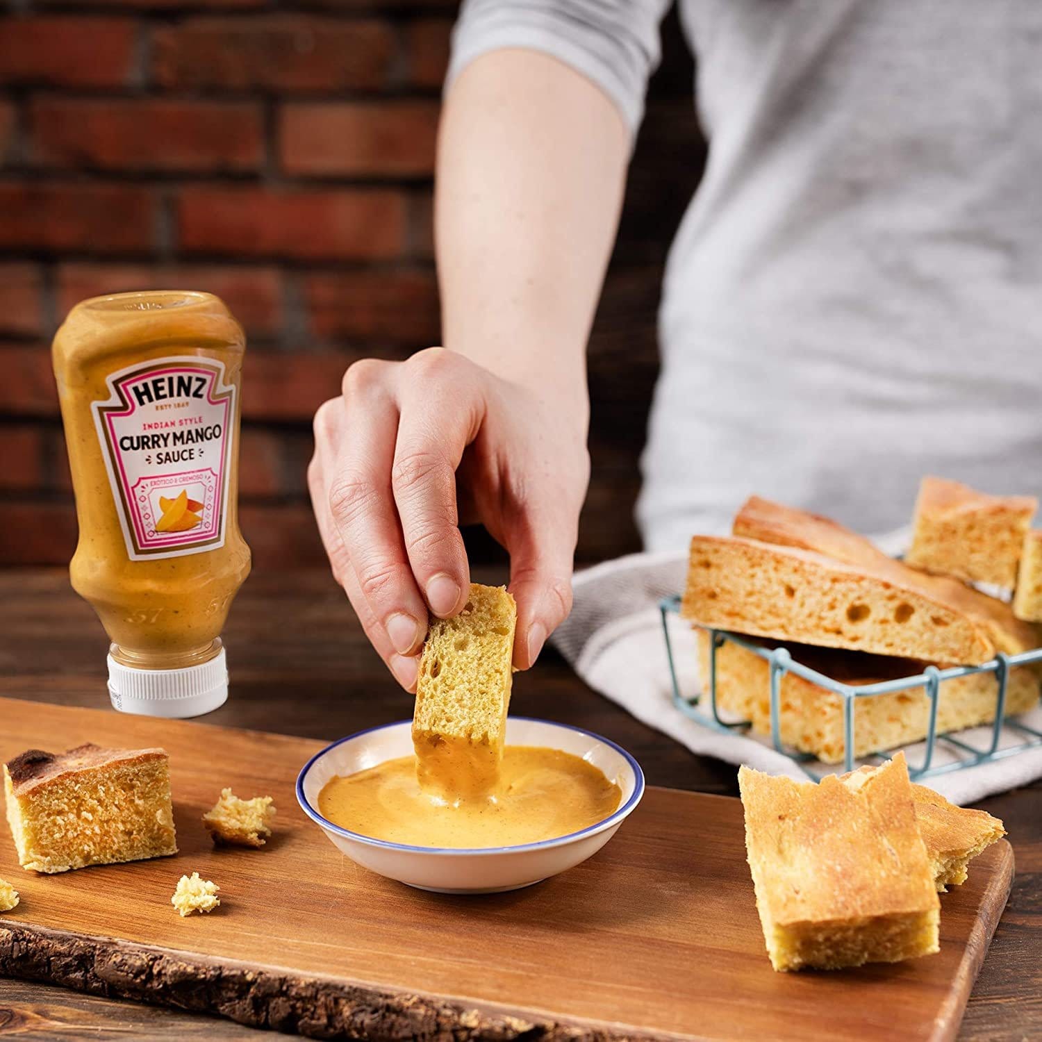 Un bol blanc avec une sauce orangée sur une planche en bois et une main qui y trempe un bout de pain. A gauche une bouteille de sauce orange Heinz et à droite un panier de pain