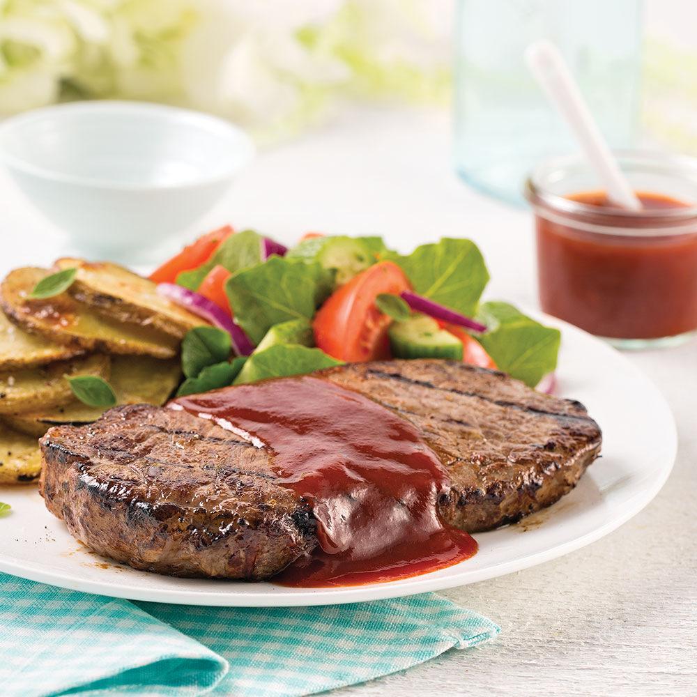 Une assiette blanche sur une table blanche avec un steak et sa sauce rouge, une salade et des pommes de terre. Derrière, à droite un bol transparent avec une sauce rouge