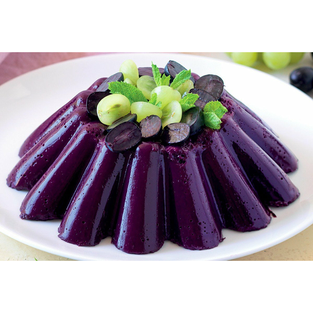 Une gelée mauve avec sur le haut des morceaux de raisins verts et noirs dans une assiette blanche