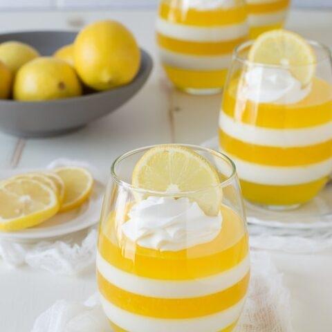Des couches de gelée jaune et de crème dans un verre transparent avec au-dessus des tranches de citron. Et à l’arrière une assiette grise remplie de plusieurs citrons