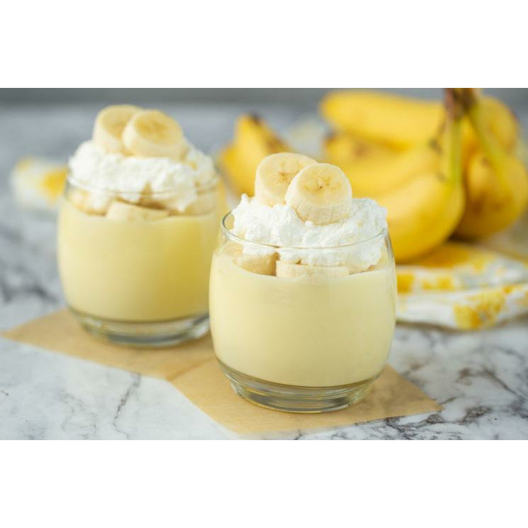 2 verres transparents remplis d’un pudding couleur crème, au-dessus de la chantilly et des morceaux de bananes. A l’arrière il y a des bananes, le tout sur une table en marbre blanc