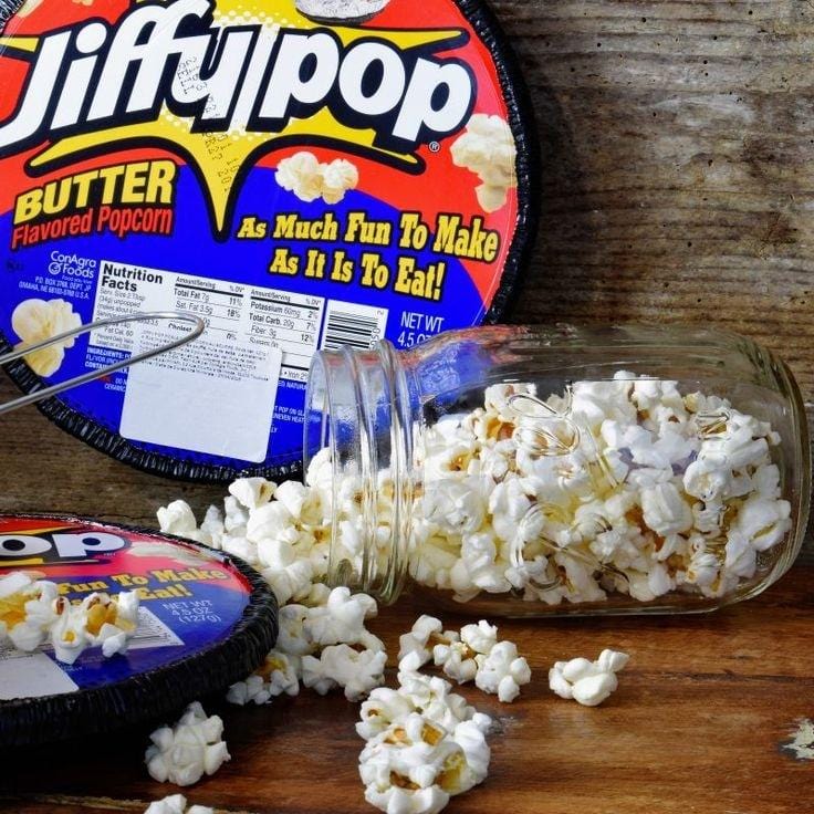 Un bocal transparent rempli de popcorn blanc qui sorte du bocal, gauche il y a un paquet rond en aluminium bleu et rouge. Le tout sur un mur et une table en bois