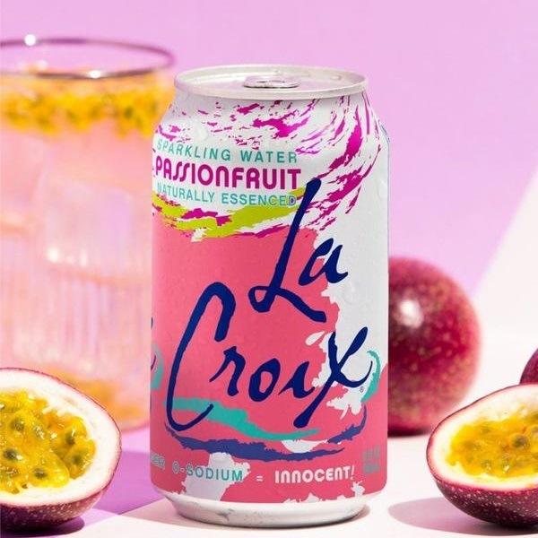 Une canette blanche et rose et des traits bleus sur une table blanche, autour des fruits de la passion. Derrière un fond rose et un verre rempli de glaçons et de fruits de la passion