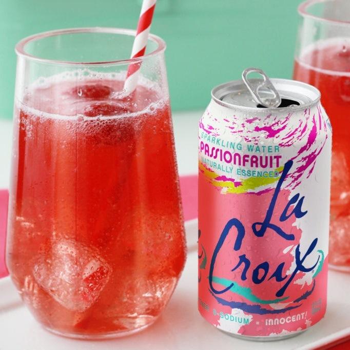 Une canette blanche et rose et des traits bleus sur une table blanche, à côté il y a un verre transparent avec une boisson rouge et des glaçons avec une paille blanche et rouge