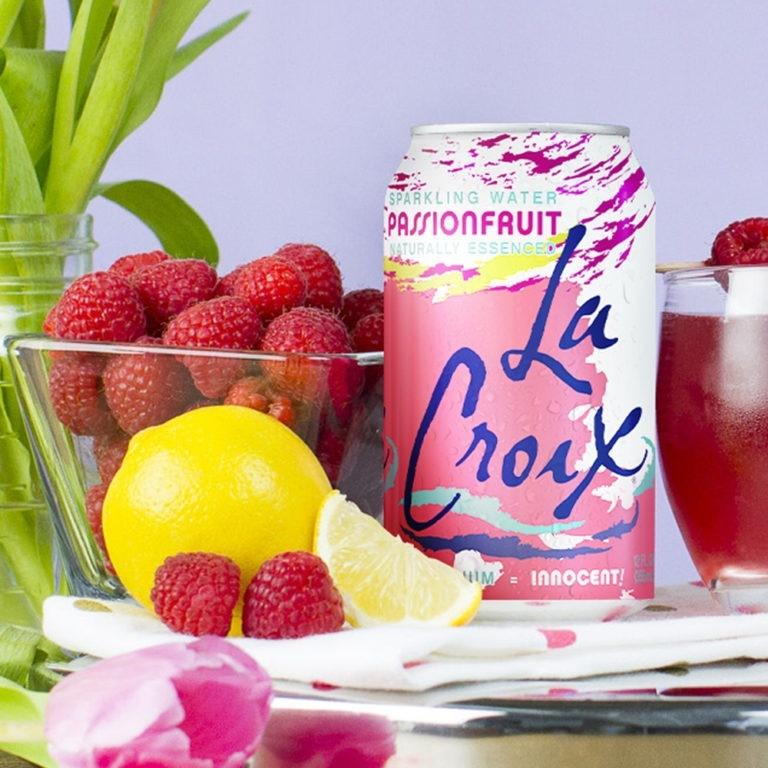 Une canette blanche et rose et des traits bleus sur une table blanche, à côté il y a un bol transparent remplie de framboises rouges et devant il y a un citron