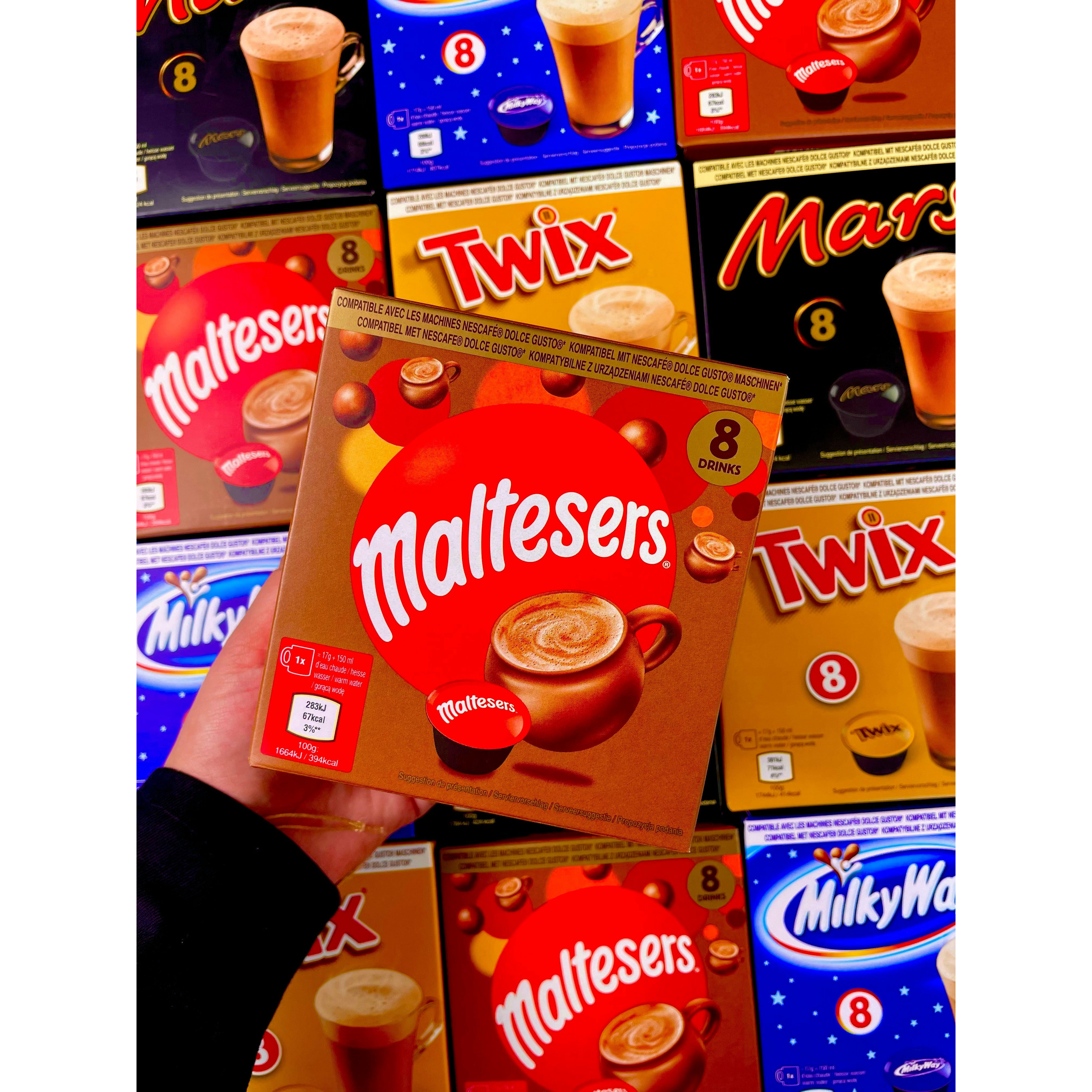 Une main tient un carton de Pods Dolce Gusto Maltesers et derrière il y a plein de cartons Twix, Maltesers, Mars et Milky Way